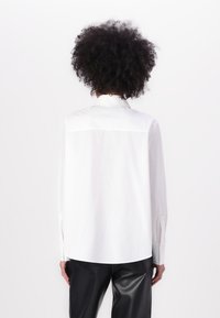 Camicia bianca a maniche lunghe con colletto a punta, dotata di yoke posteriore e polsini con bottone, abbinata a pantaloni neri lucidi e su misura.