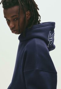 Navy blue hooded sweatshirt with large embroidered logo on the hood. Soft texture and relaxed fit. Minimalist design.