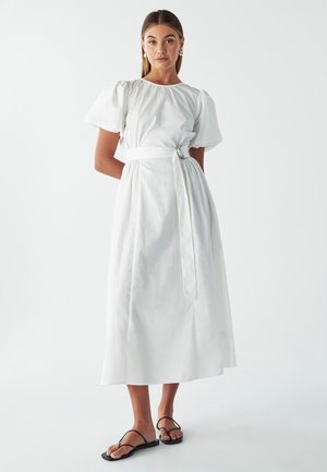 Weißes Midi-Kleid aus Baumwolle, mit Puffärmeln, tailliertem Schnitt mit gebundenem Gürtel und geradem Rock. Kombiniert mit schwarzen flachen Sandalen.