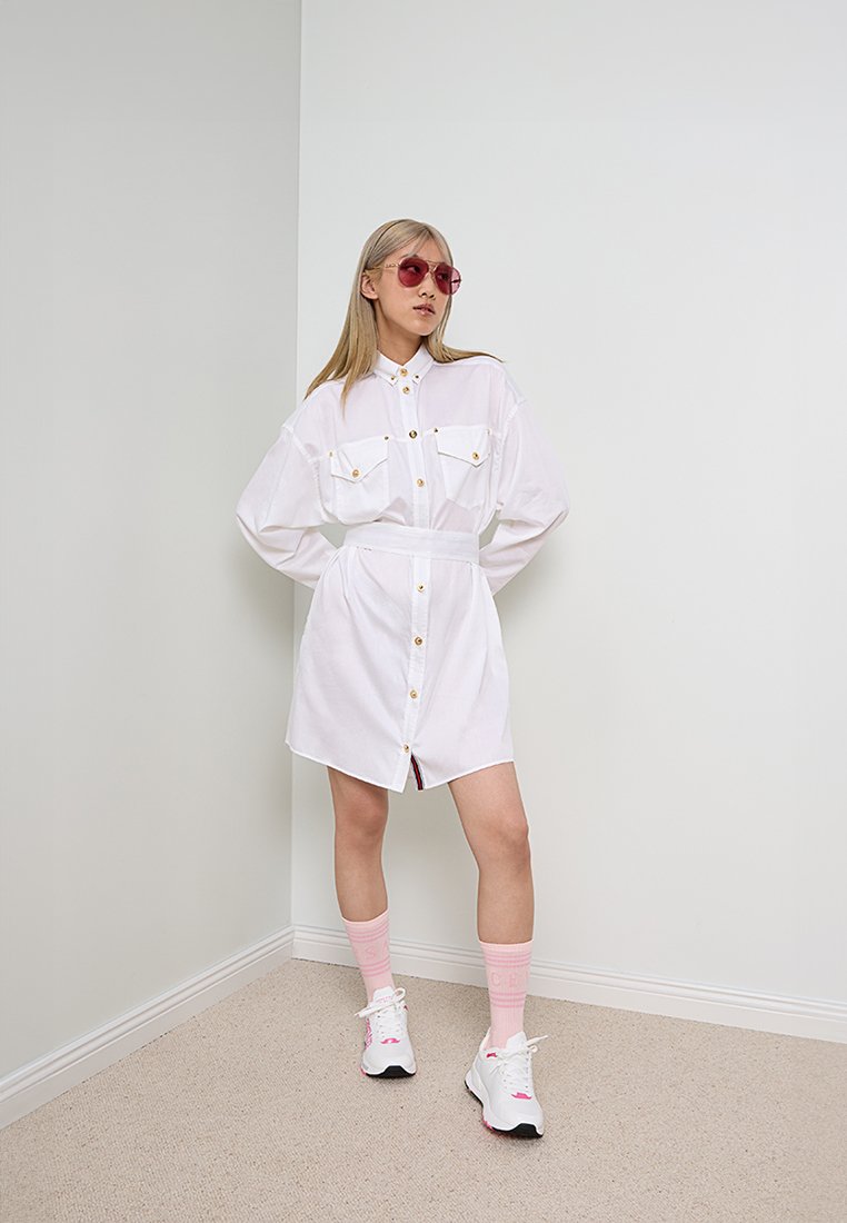 Vestido blanco de botón con dos bolsillos en el pecho, botones dorados y cintura atada. La modelo lleva gafas de sol rosas, calcetines rosas y zapatillas blancas.