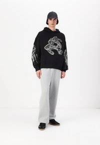 Svart hettegenser med dristig grafisk design på ermene, kombinert med grå sweatpants og svarte joggesko. Modellen har på seg en svart caps.
