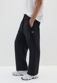 Schwarze Jogginghose mit lockerer Passform, elastischem Bund und Kordelzug. Dezentes Logo-Detail auf der Vorderseite der Tasche. Kombiniert mit grauen Turnschuhen.