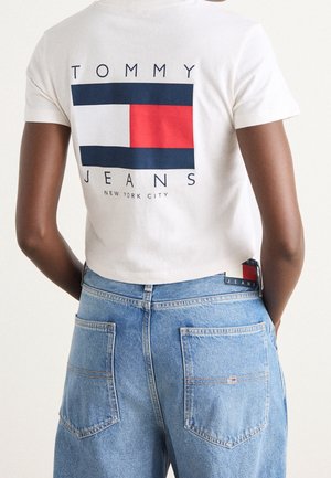 Personne portant un crop-top blanc avec un grand logo Tommy Jeans et un jean bleu, vue de dos.