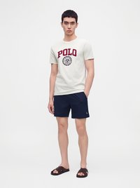 Jeune homme portant un t-shirt Polo blanc, un short bleu marine et des sandales marron, se tenant devant un fond clair uni.