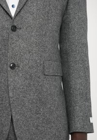 Grå tweed jakkesætjakke med en skræddersyet pasform, tre sorte knapper, en enkelt brystlomme og en kontrasterende hvid skjorte under.