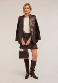 Blazer di pelle marrone, top crema, gonna a quadri, stivali al ginocchio marroni, tiene una borsa scura con accenti blu. Texture lisce, design su misura.