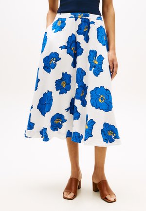 Weißer Midirock mit großem blauem Blumenmuster, getragen mit braunen Sandalen mit Absatz und einem marineblauen Oberteil, das oberhalb der Taille gekürzt ist.