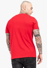 Rød bomulds T-shirt med klassisk rund halsudskæring og korte ærmer. Glat tekstur og enkel design, set bagfra. Sorte jeans synlige.