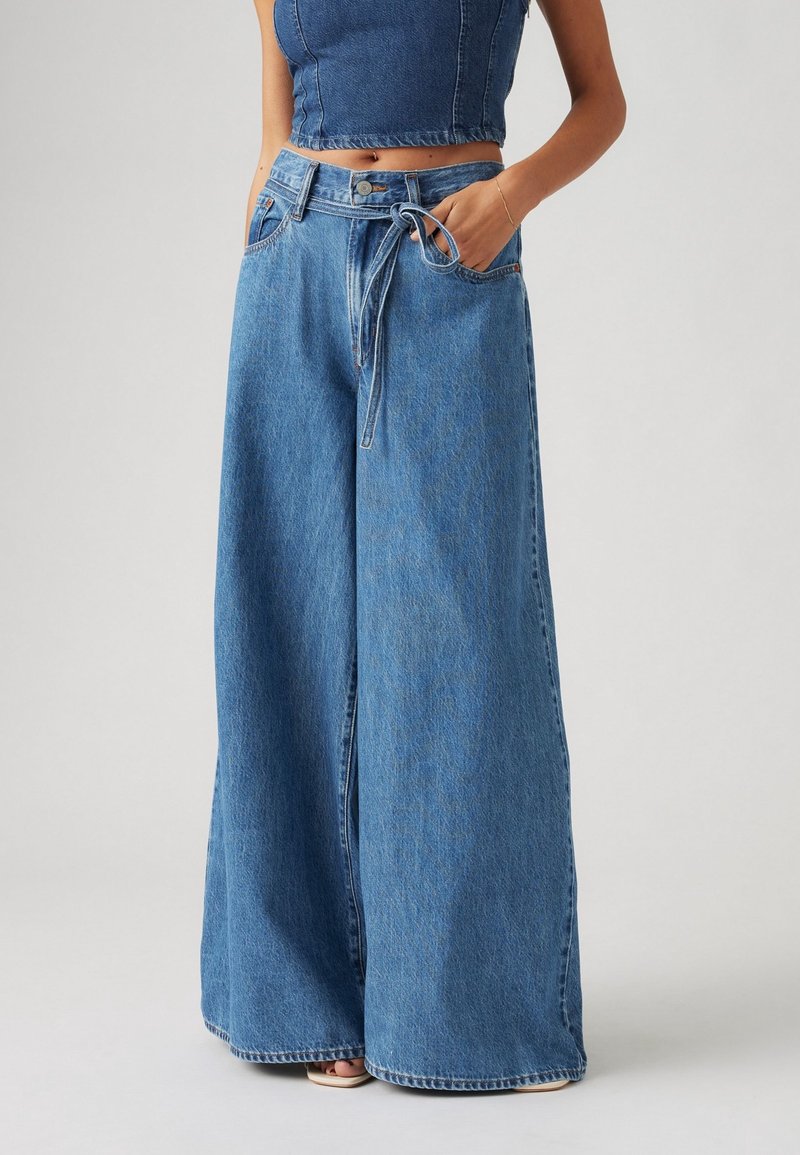 Femme portant un jean en denim bleu taille haute à jambes larges avec une ceinture, associé à un top court en denim et des sandales.