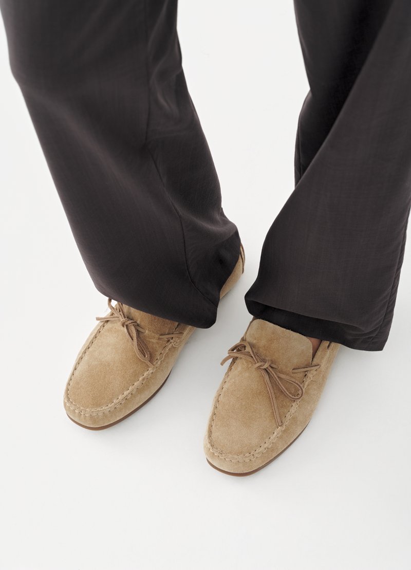 Een paar lichtbruine suède mocassinslippers gedragen bij een zwarte broek op een witte achtergrond.
