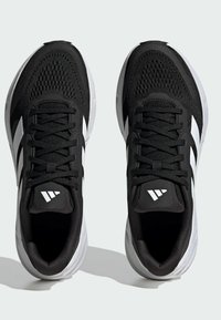 Mustat urheilukengät, joissa on hengittävä päällinen, valkoiset raidat ja logot, pyöreä kärki ja teksturoitu pohja pidon saavuttamiseksi.