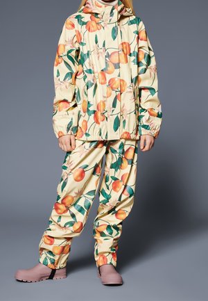 Enfant portant une combinaison de pluie crème avec un imprimé de fruits orange et de feuilles vertes, assortie à des bottes en caoutchouc roses, debout devant un fond gris.