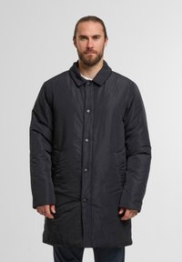 Zwarte gewatteerde jas met een kraag, sluiting met vijf knopen, zijzakken en een licht gewatteerde textuur. Draag het over een wit shirt.