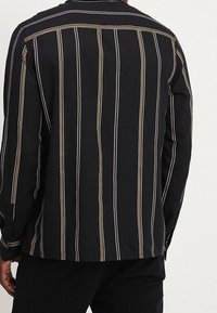 Zwarte shirt met witte en gouden verticale strepen, voorzien van een klassieke kraag, knoopmanchetten en een achterste schoudernaad detail. Zachte stoftextuur.