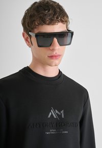 Felpa nera con logo in rilievo, abbinata a occhiali da sole neri angolari con montatura spessa e lenti scure. Texture liscia.