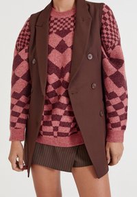 Layered outfit featuring a brown vest over a pink and maroon patterned sweater, paired with dark pinstriped shorts. Smooth textures visible.
