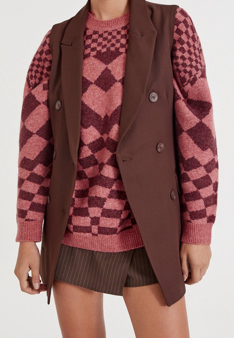 Layered outfit featuring a brown vest over a pink and maroon patterned sweater, paired with dark pinstriped shorts. Smooth textures visible.