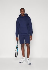 Marineblå hættetrøje med kengurulomme, parret med matchende shorts. Hvide sneakers og et tennisrack er inkluderet i outfittet.