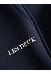 Tejido azul marino con una textura suave y un logo blanco de "LES DEUX". El diseño enfatiza la simplicidad y el estilo moderno en la vestimenta.