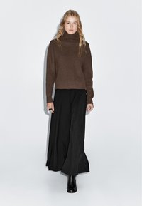 Maglione a collo alto lavorato a maglia marrone con texture a coste, abbinato a una gonna nera fluida e stivaletti neri, esposto su uno sfondo chiaro.