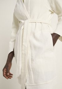 White textured robe featuring a belt, two front pockets, and wide sleeves. Made from quilted fabric with a soft, lightweight appearance.