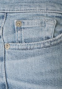 Tejido de mezclilla azul claro con un primer plano de un bolsillo trasero, que presenta costuras visibles y dos remaches metálicos en los bordes del bolsillo.