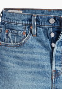 Jean en denim bleu en gros plan montrant la braguette à boutons, la boucle de ceinture, les rivets et un petit détail usé sur la poche avant.