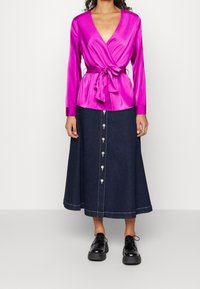 Top avvolgente in satin fucsia con maniche lunghe e dettaglio con fiocco abbinato a una gonna in denim scuro con bottoni, caratterizzata da cuciture bianche e una silhouette svasata.