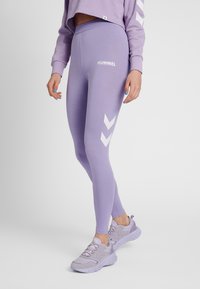 Vrouw met lichtpaarse Hummel legging en bijpassende sneakers, staand met één been iets naar voren tegen een effen achtergrond.