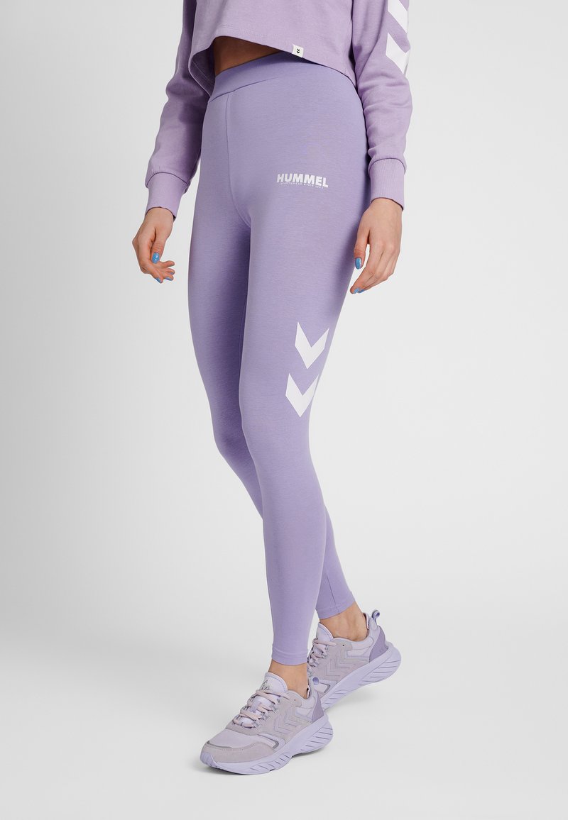 Vrouw met lichtpaarse Hummel legging en bijpassende sneakers, staand met één been iets naar voren tegen een effen achtergrond.