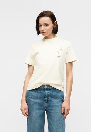 Tânără purtând un tricou crem cu mâneci scurte, cu design de pahar de martini și blugi albaștri, stând pe un fundal simplu și deschis la culoare.