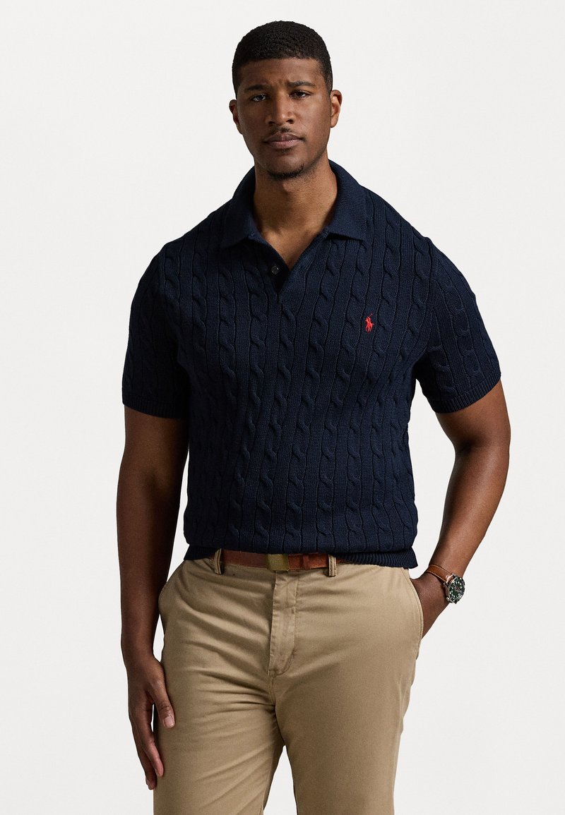 Μπλέ πόλο κοντομάνικο πλεκτό με κολάρο και κόκκινο λογότυπο. Συνδυασμένο με καφέ παντελόνι, που διαθέτει καφέ ζώνη και ρολόι χειρός.