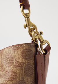 Coin d'un sac en cuir marron et toile avec fermoir doré et étiquette en cuir attachée à une chaîne de perles dorées sur fond blanc.