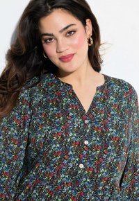 Blomstret bluse med sort baggrund, røde og blå blomster mønstre, V-hals og sølvknapper ved halsen. Glat stofstruktur.