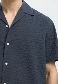 Kortsleppet, mørkeblå knappet skjorte med teksturert rutenettmønster, med spiss krage og lyse knapper.