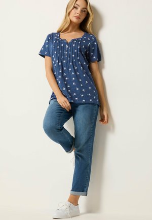 Femme appuyée contre un mur, portant une chemise bleu marine à manches courtes avec des imprimés de palmiers blancs, un jean bleu et des baskets blanches.