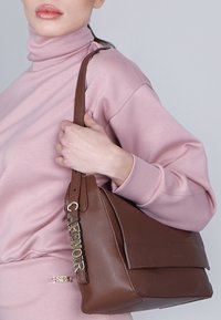 Bruine leren handtas met een zachte textuur en een gebogen vorm. Beschikt over een opvallend metalen detail met de tekst "CAFÉ NOIR" op de strap.