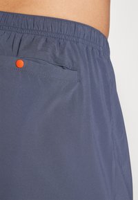 Navy blue shorts with an elastic waistband, featuring a rear pocket with an orange button, and a smooth, lightweight fabric texture.