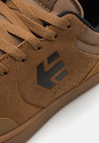 Καφέ σουέτ sneakers με μαύρες λεπτομέρειες, διαθέτοντας υφασμάτινα πάνελ με υφή, επίπεδες κορδόνια και λογότυπο στο πλάι. Λαστιχένια σόλα με τρυπήματα.