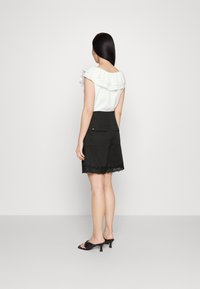 Weiße Bluse mit Rüschenausschnitt, kombiniert mit schwarzen Shorts mit Spitzensaum und Gesäßtaschen. Das Modell trägt schwarze High-Heelsandalen.