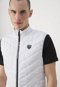 Bijela quilted prsluk s visokim ovratnikom, s crnim patentnim zatvaračem i izvezenim logom. Nosite preko crne majice kratkih rukava. Glatka tekstura.