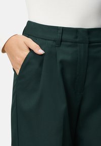 Pantalons ajustés verts en tissu lisse, avec un devant plissé et des poches latérales, dotés d'une taille mi-haute.