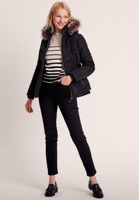 Schwarze Puffjacke mit einem mit Futter aus Pelz gefütterten Kapuzen, kombiniert mit einem gestreiften Strickpullover, schwarzen Hosen und glänzenden Slippern.