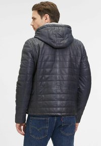 Giacca di pelle nera con cappuccio, texture trapuntata, design attillato e lucentezza sottile. Indossata con jeans blu chiaro, cuciture posteriori visibili.