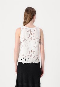 Hvit ermeløs topp laget av blonder med blomstermønster, med rund hals og åpen ryggdesign, kombinert med et svart strukturert skjørt.