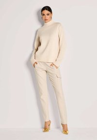 Frau in einem cremefarbenen Rollkragenpullover und beigen Cargo-Hosen, Hände in den Taschen, trägt goldene High Heels vor einem hellen Hintergrund.