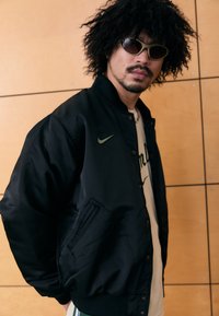 Musta bomber-takki, jossa on sileä pinta ja Nike-logo vasemmassa rinnassa. Mallilla on vaaleat aurinkolasit ja valkoinen paita.