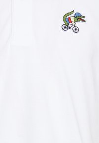 Bílá polo košile vyrobená z texturované tkaniny, která má vyšitý design zeleného krokodýla na bicyklu s modrou přilbou a třemi barvami na těle.