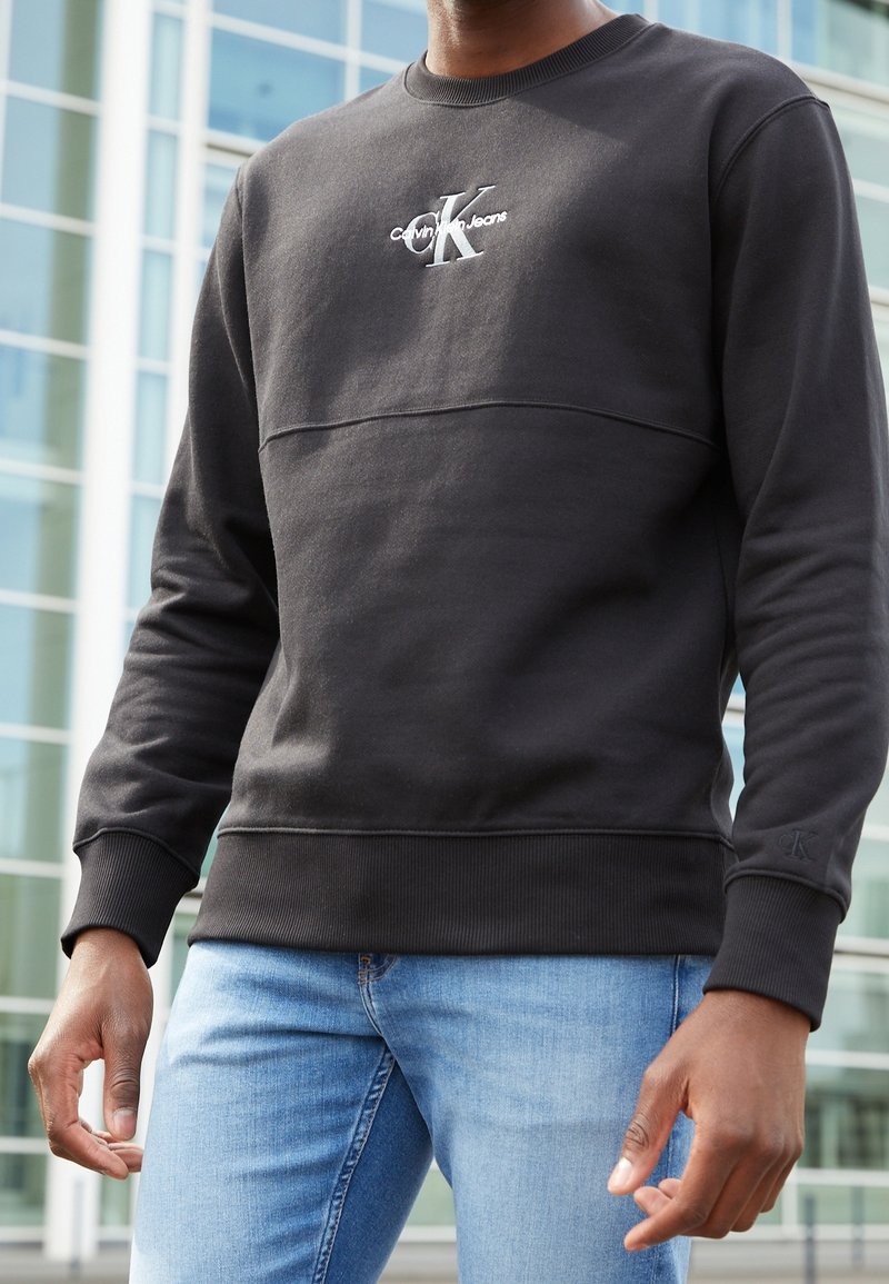 Svart sweatshirt med strukturerad yta, utrustad med rund halsringning och en Calvin Klein Jeans-logga på bröstet. Ribbstickade muddar och fåll.