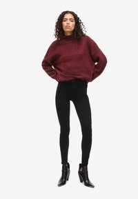 Burgundi csíkozott garbó pulóver laza szabásban, fekete skinny farmernadrággal és fekete, magas sarkú bokacsizmával párosítva, fehér háttér előtt.